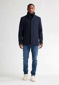 Laivastonsininen villasekoitetakki korkealla kauluksella, vetoketjulla edessä ja kahdella sivutaskulla, yhdistettynä sinisiin farkkuihin ja beigeihin lenkkareihin.