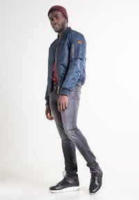 Veste aviateur bleu marine avec fermeture éclair et poche latérale, associée à un jean foncé et des baskets noires ; le mannequin porte un bonnet bordeaux.