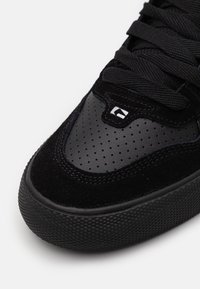 Zapatilla negra que combina ante y cuero perforado, con punta redonda, suela de goma negra texturizada y gruesa, y cordones planos negros.