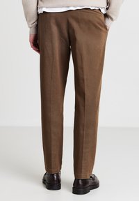 Mann trägt braune gerade geschnittene Hosen und dunkelbraune Lederdressschuhe, steht mit den Händen in den Taschen vor einem weißen Hintergrund.