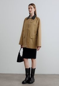 Ταμπά εξωτερικό μπουφάν με μαύρο γιακά, φερμουάρ μπροστά και δύο τσέπες επιδιορθώσεων. Φορεμένο πάνω από μαύρη φούστα και συνδυασμένο με μαύρα μποτάκια αστραγάλου.