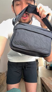 Jeune homme en chemise blanche et short noir tient un sac bandoulière noir avec le logo "BOSS" dans le salon.