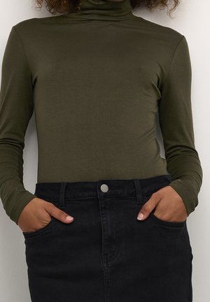 Henkilö, jolla on päällään oliivinvihreä pitkähihainen poolopaita, joka on sullottu korkeavyötäröisen mustan denimhameen sisään, kädet taskuissa.