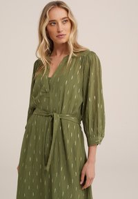Rochie verde cu mâneci lungi, decolteu în V, legată la talie, cu accente aurii decorative. Material texturat, ușor, cu o silueta fluidă.