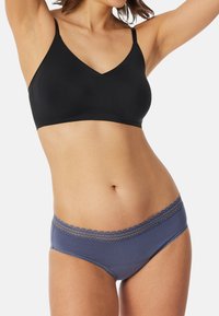 Frau in schwarzem nahtlosem BH und blauen Hüftslip-Unterhosen mit Spitze, die mit erhobenen Armen vor einem schlichten Hintergrund posiert.