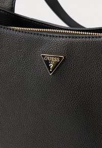 Melna teksturēta somiņa ar zelta metāla detaļām un trīsstūra formā Guess logo. Rāvējslēdzēja aizdare augšpusē, izgatavota no ādas materiāla.