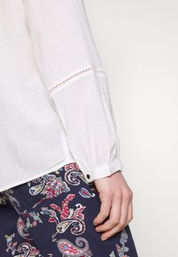 Blouse blanche à manches longues avec tissu texturé et bordure en dentelle aux poignets, associée à un pantalon bleu orné d'un motif cachemire vibrant.