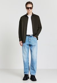 Camicia verde scuro con bottoni, t-shirt bianca, jeans a gamba larga di colore azzurro chiaro e scarpe nere. Gli accessori includono occhiali da sole quadrati e una cintura nera.