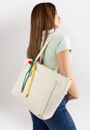Donna con capelli lunghi castani che indossa una maglietta verde chiaro e jeans blu, che porta una grande borsa a tracolla crema con un ciondolo intrecciato colorato.