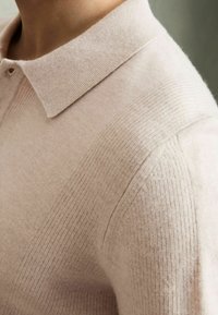 Polo en tricot rose clair avec une texture côtelée et un col classique. Présente de subtiles rayures horizontales et une apparence douce et confortable.