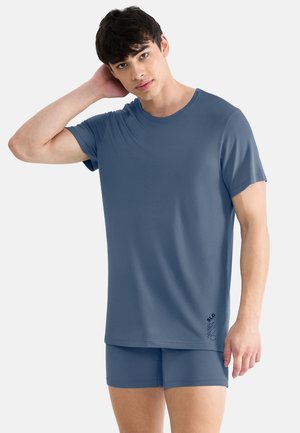 Young man with short dark hair wearing a blue short-sleeve shirt and matching shorts, posing with one hand behind his head.