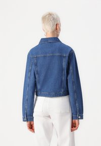 Jaqueta de ganga azul cortada com mangas longas, punhos com botões e colarinho clássico. Apresenta costuras visíveis e uma textura ligeiramente desbotada.