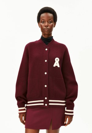 Frau trägt eine burgunderrote Collegejacke mit weißen Knöpfen und Streifen, mit einem weißen Buchstaben A auf der Brust, kombiniert mit einem passenden Rock.