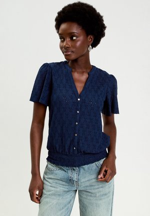 Granatowa bluzka z krótkim rękawem o koronkowej fakturze, z dekoltem w serek, zapinana na guziki z marszczonym dołem. Połączona z jasnoniebieskim dżinsem.