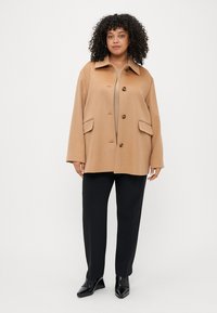 Κάμελ χρωματιστό oversized παλτό με γιακά, μπροστινά κουμπιά και πλαϊνές τσέπες, σε συνδυασμό με μαύρο παντελόνι και γυαλιστερά παπούτσια. Ίσια υφή υφάσματος.