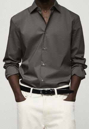 Ciemnoszara, długorękawowa koszula na guziki z podwiniętymi rękawami, zestawiona z kremowymi spodniami i czarnym paskiem z srebrną klamrą. Ręce w kieszeniach.