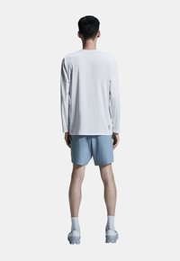 Langærmet hvid skjorte, lyseblå shorts, står på en ensfarvet baggrund. Har logo på skjorten og struktureret fodtøj.