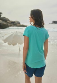 T-shirt turquoise à manches courtes en tissu doux, associé à un short en denim sombre. L'arrière-plan présente une plage, de l'eau et un parasol.