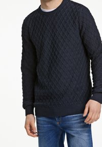 Marineblauer Strickpullover mit einem strukturierten Rautenmuster, rundem Ausschnitt und gerippten Bündchen und Saum, kombiniert mit blauen Jeans.