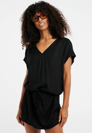 Zwart kort jurkje met een V-hals, korte mouwen, elastische taille met trekkoord en een gladde textuur. Model draagt een oversized bruin zonnebril.