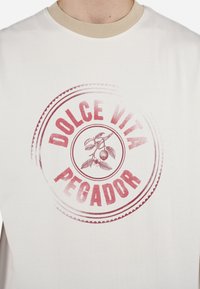 Balta kokvilnas T-kreklā ar bēšas krāsas ribotu apkakli, ar apaļu vintage stila grafiku sarkanā krāsā, kurā rakstīts "DOLCE VITA PEGADOR" un ķirši.