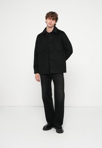 Sort jakke med to brystlommer, lavet af et tykt stof, med knapper foran og en klassisk krave. Kombineret med sorte bukser og sko.