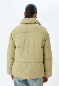 Giacca imbottita color verde oliva chiaro, con colletto alto, texture trapuntata e vestibilità ampia. Abbinata a jeans in denim azzurro.