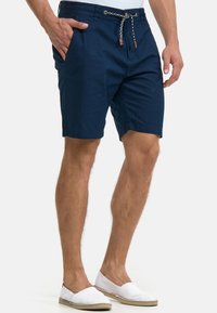 Shorts en coton bleu marine avec taille à cordon de serrage comprenant des accents tissés, des poches latérales et une longueur mi-cuisse. Portés avec des chaussures blanches à enfiler.