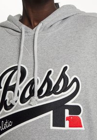 Gray hoodie made of soft fabric, featuring a large black embroidered logo, drawstrings, and a white outline. Accents include a red logo detail.