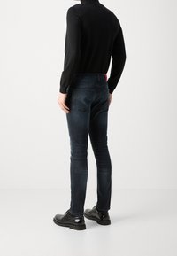 Jean en denim foncé avec une coupe slim et une décoloration minimale. Comporte des poches arrière et un petit accent rouge sur la ceinture. Associé à des chaussures noires.