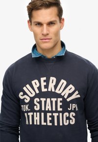 Felpa navy con collo rotondo, maniche lunghe e una stampa grafica cream ben visibile che riporta la scritta "SUPERDRY STATE JAPAN ATHLETICS".