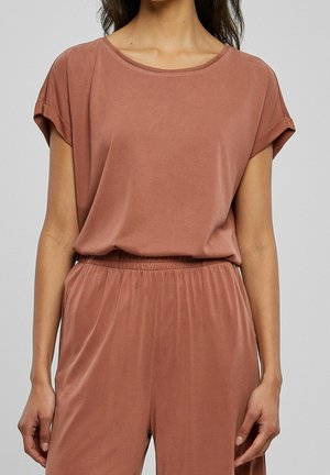 Mujer que lleva una blusa holgada de manga corta de color rosa polvo y pantalones a juego con cintura elástica, frente a un fondo gris uniforme.