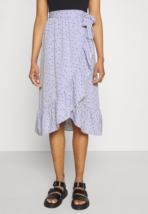 Hellblau-lavendelfarbener Wickelrock mit einer gerafften Saumkante, kleinem floralen Muster und seitlichem Bindeband. Kombiniert mit schwarzen, trägerlosen Sandalen.
