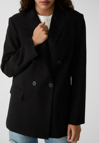 Black double-breasted blazer made of structured fabric. Features two front pockets, black buttons, and a notched lapel design.
