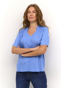 Haut bleu clair à manches courtes avec un décolleté en V et des détails froncés aux épaules. Tissu doux avec une texture lisse, associé à un jean foncé.