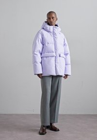 Puffy lilac down jacket with a high neck and hood, featuring front pockets and a boxy silhouette, paired with gray wide-leg trousers.