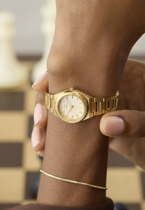 Primer plano de una mano ajustando un reloj de pulsera Timex dorado con una esfera de patrón claro, sobre un fondo desenfocado de un tablero de ajedrez.