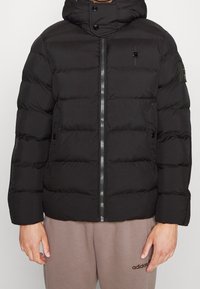 Černá péřová bunda se zipem vpředu, kapucí a prošívanými panely. Vlastnosti zahrnují logo na hrudi a rameni, s hladkou a jemnou texturou.