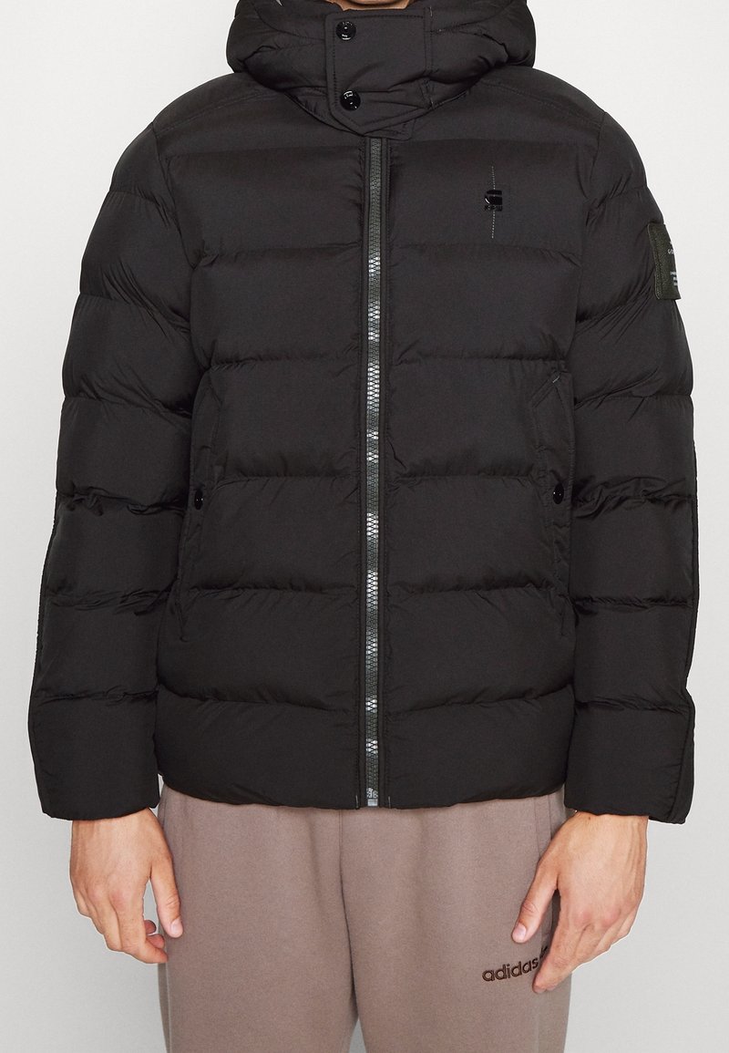 Černá péřová bunda se zipem vpředu, kapucí a prošívanými panely. Vlastnosti zahrnují logo na hrudi a rameni, s hladkou a jemnou texturou.