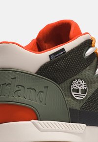 Gros plan sur une basket Timberland verte et orange avec tissu texturé, col rembourré, logo arbre en métal et étiquette imperméable TimberDry.