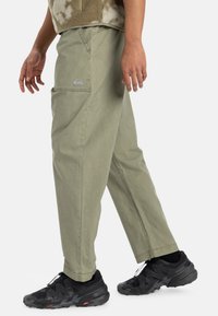 Light olive green cargo pants with a straight cut, featuring a logo on the side, pockets, and a sturdy cotton fabric. Black athletic shoes complete the look.