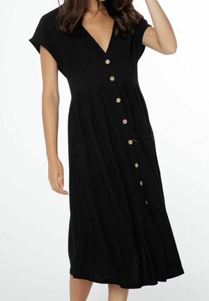 Woman wearing a black midi dress with short sleeves and beige buttons down the front, standing against a white background.