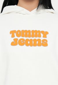 Felpa bianca con cappuccio caratterizzata da un ampio logo arancione "TOMMY JEANS". Il materiale sembra morbido; il design è casual con una vestibilità rilassata e cappuccio.