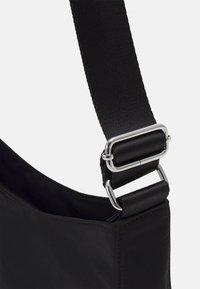 Černá látková taška s hladkou texturou, která má široký řemínek a stříbrné kovové spony. Horní část je otevřená s minimálním prošíváním.