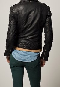 Veste en cuir noir avec des accents matelassés, des poignets zippés et un ourlet à côtes. Portée par-dessus une chemise bleu clair et un pantalon vert foncé.