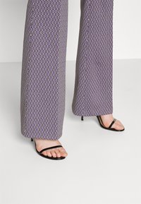 Jambes portant un pantalon large à motifs violets avec des sandales à talons hauts ouvertes noires sur un sol gris clair.