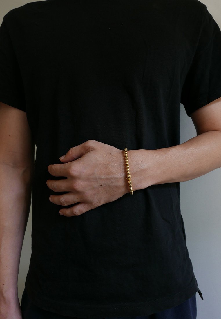 Brăţară din mărgele de aur purtată pe o încheietură, având mărgele rotunde, lucioase. Purtaşul are un tricou de culoare neutră.