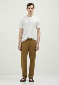 Maglietta henley bianca a maniche corte con tre bottoni, abbinata a pantaloni jogger marroni con cordino e sandali marroni. Silhouette semplice e rilassata.