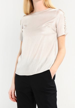 Nainen, joka pukeutunut kiiltävään beigeen lyhythihaiseen puseroon, jossa on rypytetyt yksityiskohdat hihoissa, sekä mustiin housuihin, kädet taskuissa, neutraali tausta.