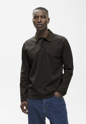 Uomo con capelli corti che indossa una camicia a maniche lunghe a righe marrone scuro e jeans blu, in piedi con una mano in tasca davanti a uno sfondo bianco.
